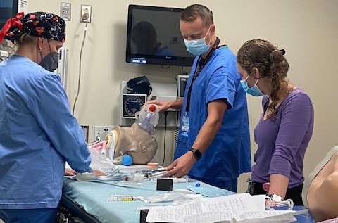 Ed Stene, AGACNP, practices airway skills at a table the simulation center with Katherine Pavlos, PA-C, looking on at right and Jennifer King Wilson, AGACNP, CRNA, at left on the other side of the table.