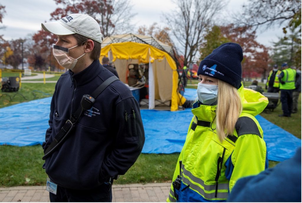 Two trainees wearing face masks watch during a training exercise, with a tent-like shelter in the background.