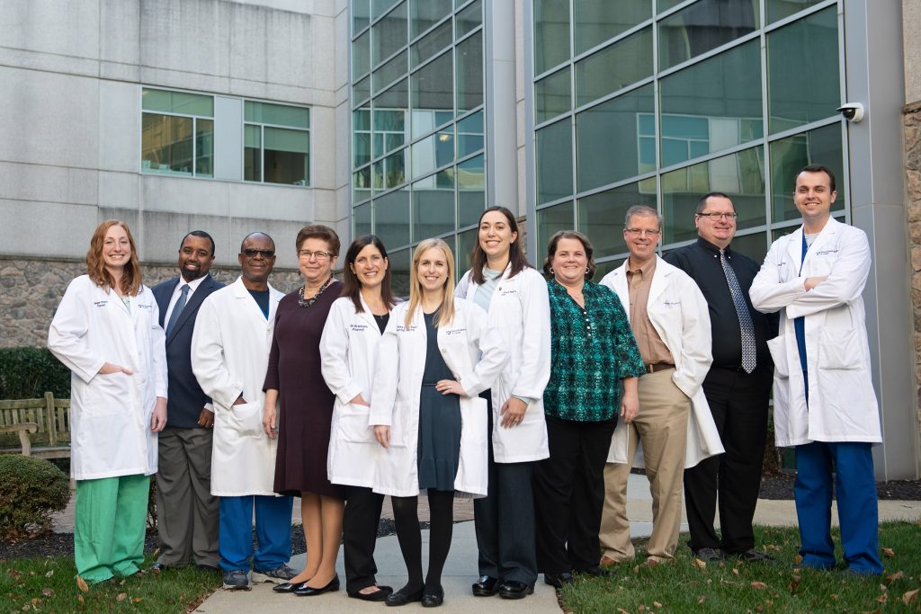 A group of 12 preceptors, some wearing white coats, stand in a line for a photo in an outdoor area with St. Joseph Medical Center in the background.