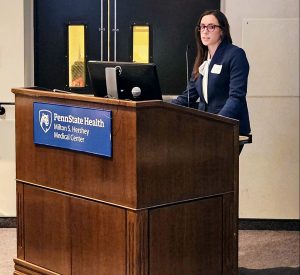 Andrea Hiller stands behind a lectern that has a computer monitor and a sign on the front with the Penn State Health Milton S. Hershey Medical Center logo
