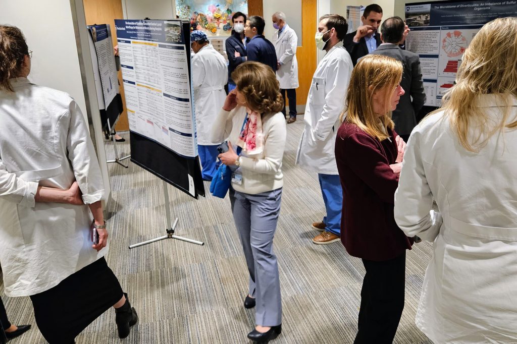 People, some in white coats and some wearing face masks, talk in small groups and look at large poster presentations