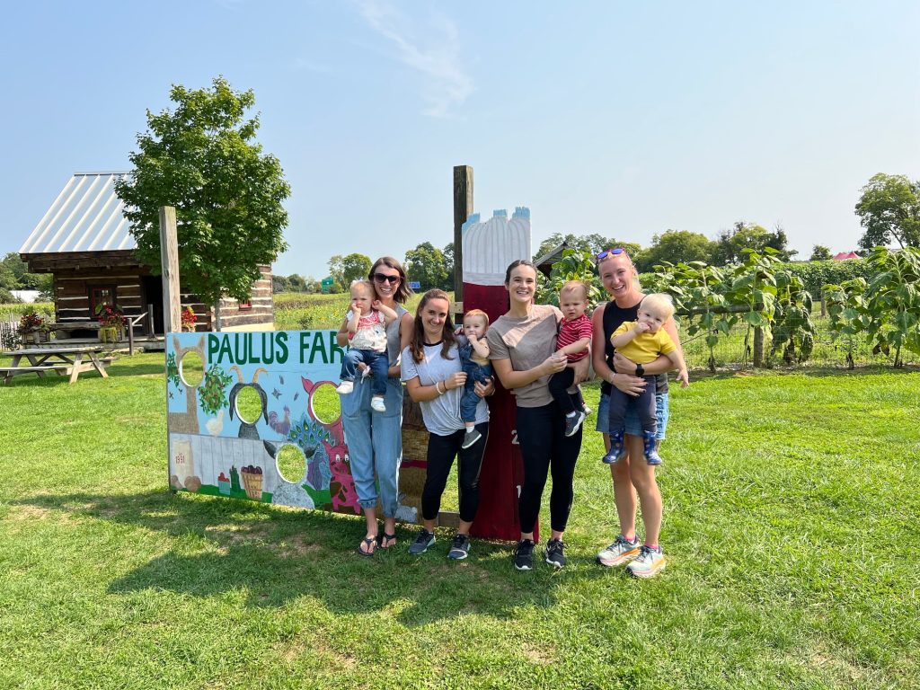 Group of 4 women, each holding a young child, standing in the right front of a Paulus Farms sign with crops and a log cabin in the background 
