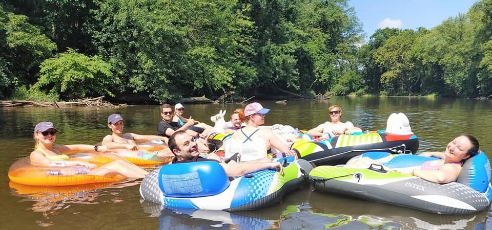 Nine people in multicolored rafts float in a tree-lined creek in summer 2022