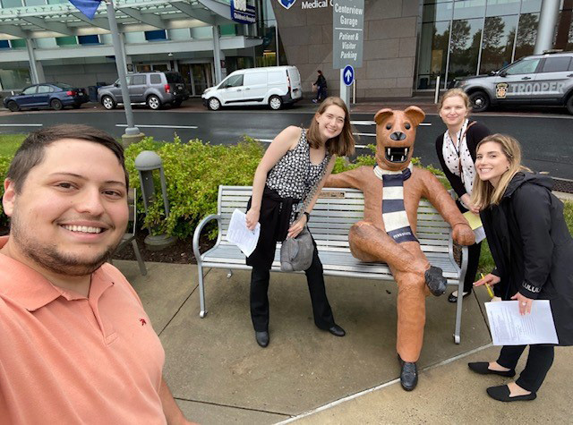 Three women pose around a Nittany Lion statue on a bench with a man in the foreground who is taking the photo.