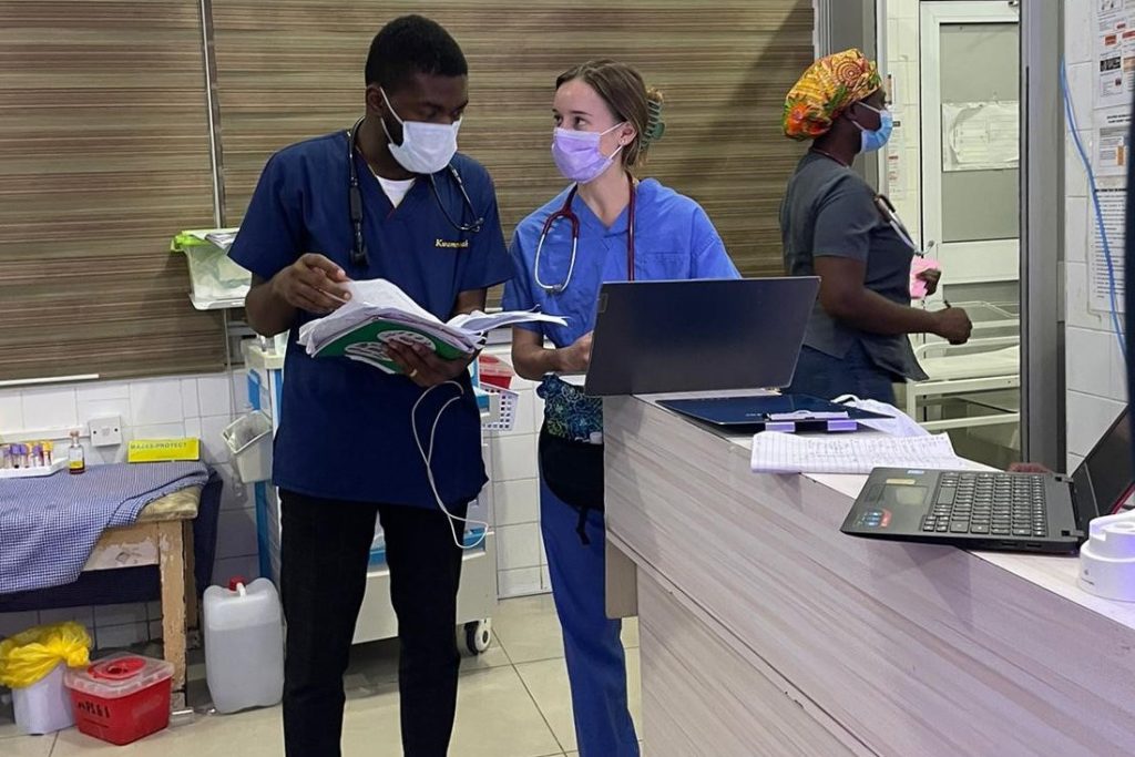 Two people in doctors' scrubs and face masks talk to each other while holding papers and with a laptop open in front of them.