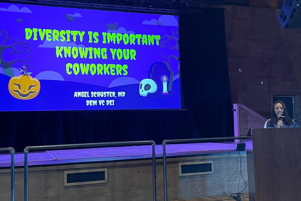 A woman talks at a lecturn; to her right a large screen shows a title slide that states Diversity is Important, Knowing Your Coworkers, Angel Schuster, MD, DEM VC DEI.