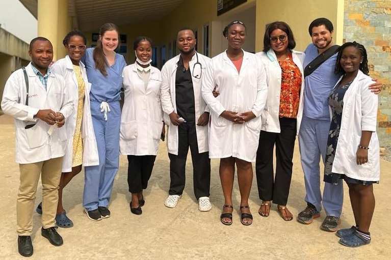 Emily Gibbons and Jake Beerel, wearing medical scrubs, pose in a line with seven other people who are all wearing white coats.