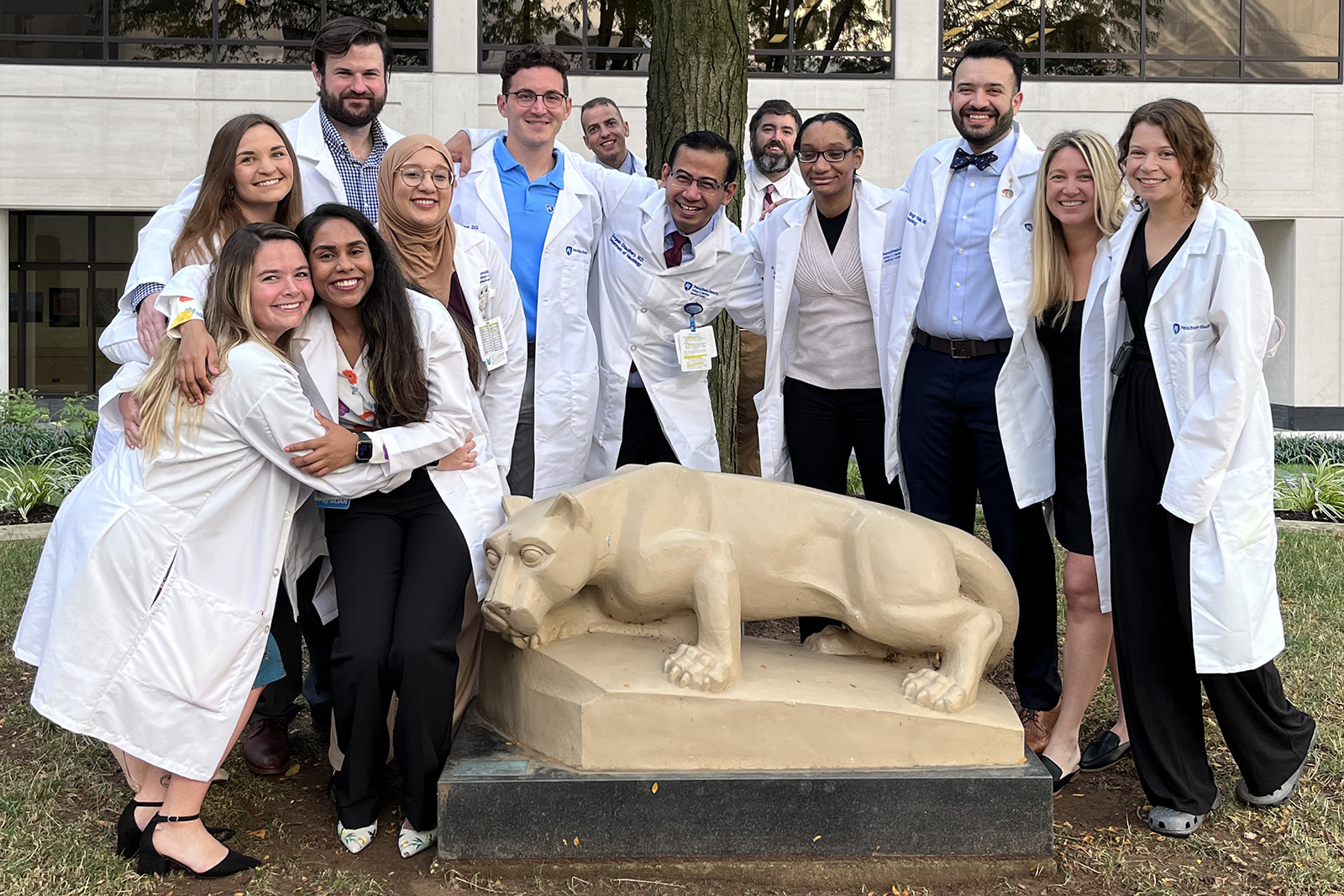 A group of 13 neurology residents, some with arms around each other and all wearing white coats, stand around a Nittany Lion statue in the Penn State College of Medicine courtyard.