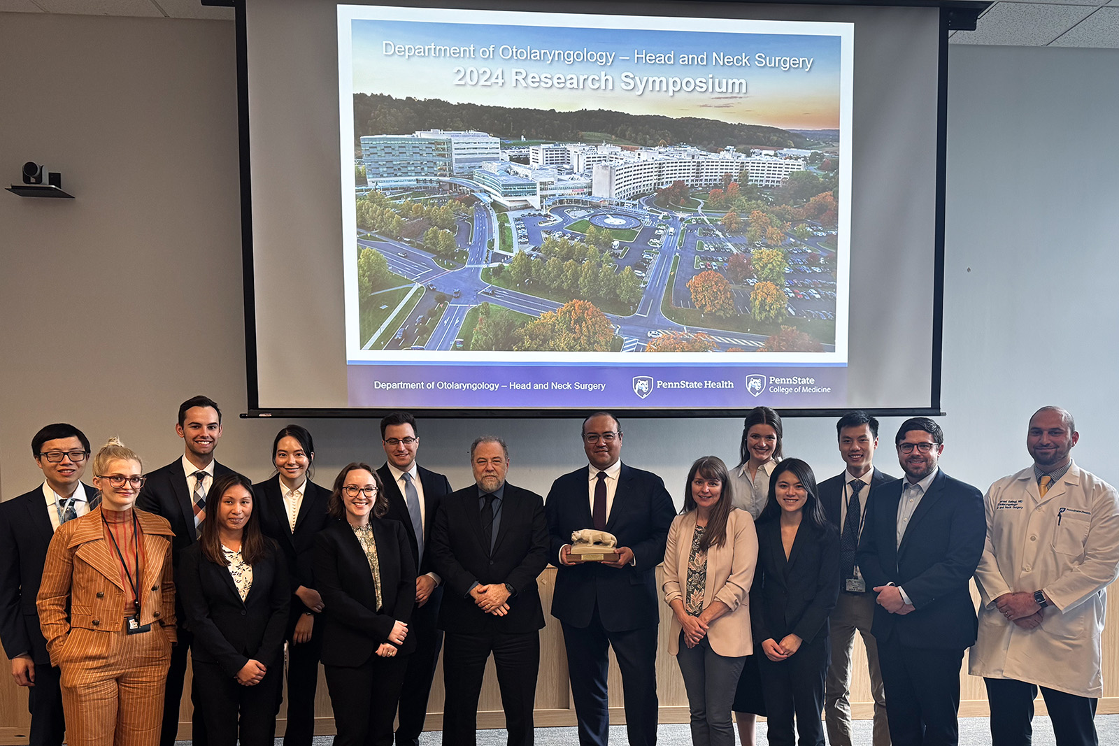 Residents and faculty stand in front of a screen with a title slide for the research symposium; Dr. Jose Zevallos is in the middle holding a Nittany Lion statue.