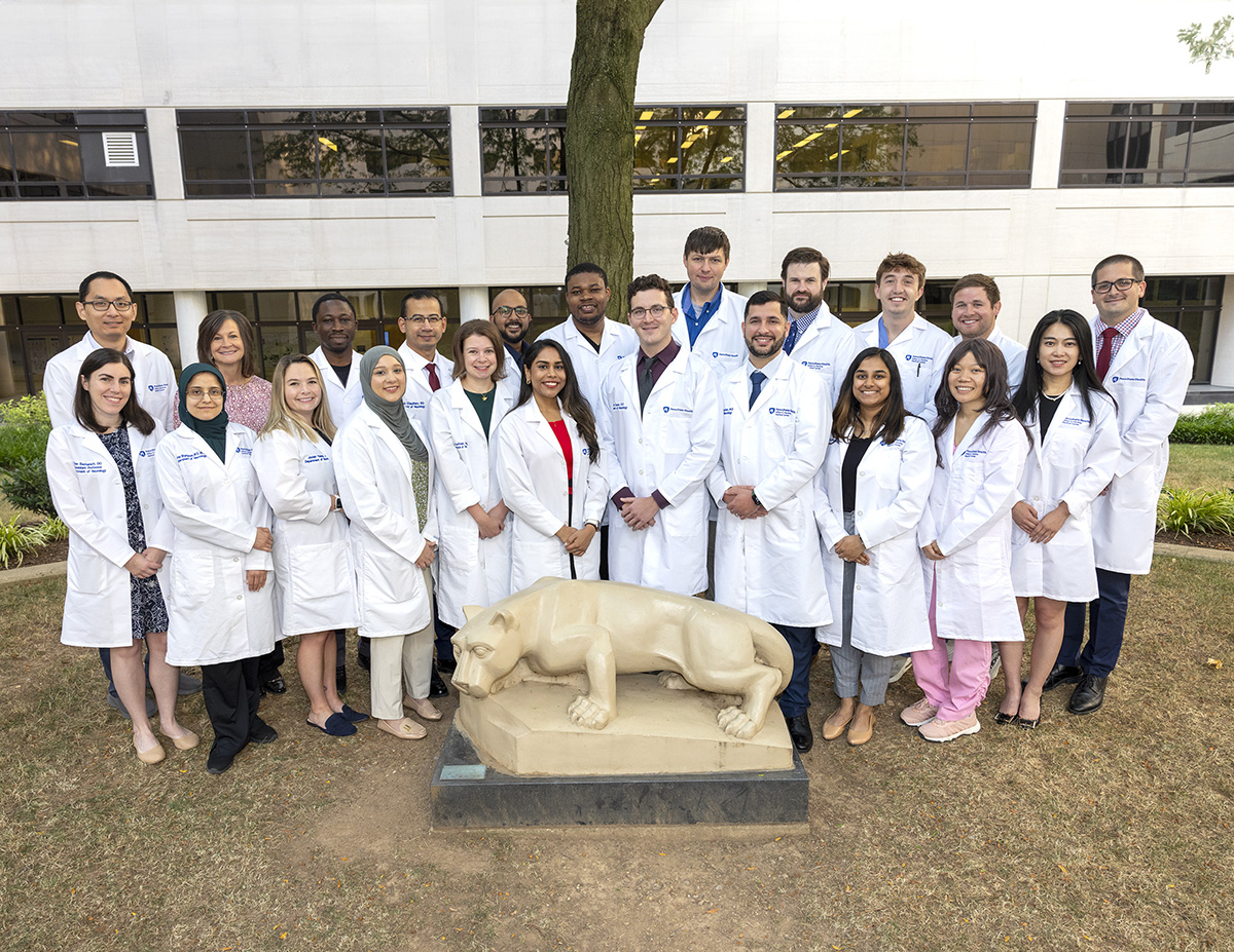 A group of residents and residency leaders wearing white coats standing behind a Nittany Lion statue in the College of Medicine courtyard