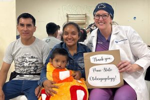 Dr. Jessyka Lighthall sits with a mother, father and child in Peru while holding a sign that says Thank you, Penn State Otolaryngology!